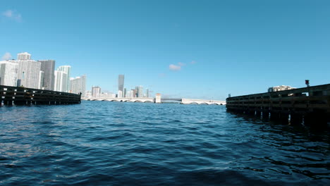 Disparo-Del-Nivel-Del-Agua-Desde-Un-Bote-Pequeño-Que-Pasa-Por-Debajo-De-Un-Puente-Con-Miami-Al-Fondo