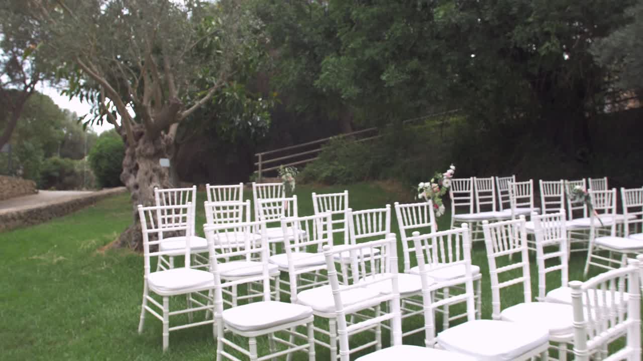 Lento movimento della telecamera lungo file di sedie bianche disposte per una cerimonia di matrimonio all'aperto su erba verde a Maiorca.