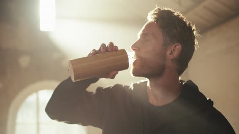 a sweaty man in a black sports uniform after a workout drinks from a special bottle in a sunny gym