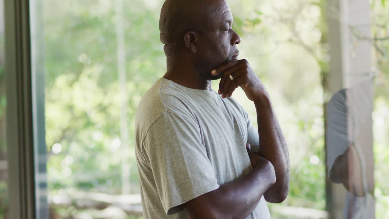 Thoughtful african american senior man standing in living room holding chin, looking out of window