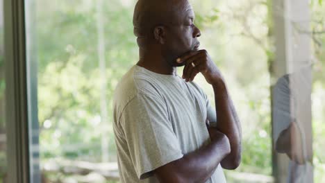 thoughtful african american senior man standing in living room holding chin, looking out of window