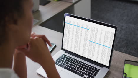mujer biracial sentada en el escritorio viendo el procesamiento de datos de codificación en la pantalla de la computadora portátil