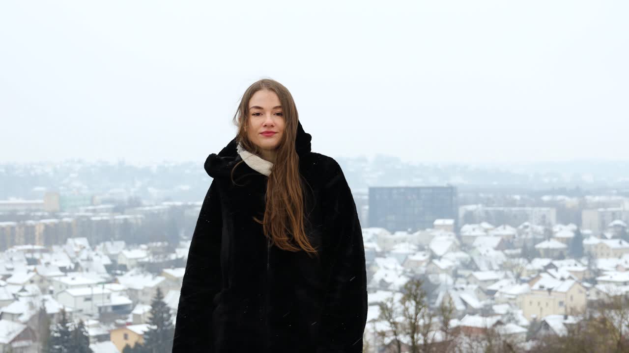 눈이 내리는 동안 검은 코트를 입고 도시 앞에서 포즈를 취하는 젊은 백인 여성 모델