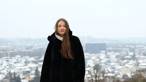 Joven-Modelo-Blanca-Posando-Frente-A-Una-Ciudad-Con-Un-Abrigo-Negro-Mientras-Nieva