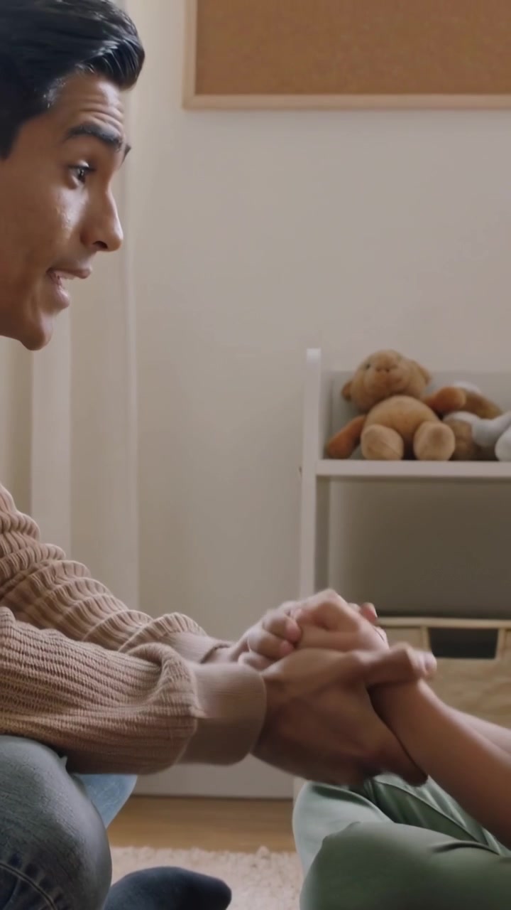 Video vertical: Hombre acercándose, sosteniendo las manos de un niño y consolándolo en un dormitorio, con suéter y un osito de peluche.