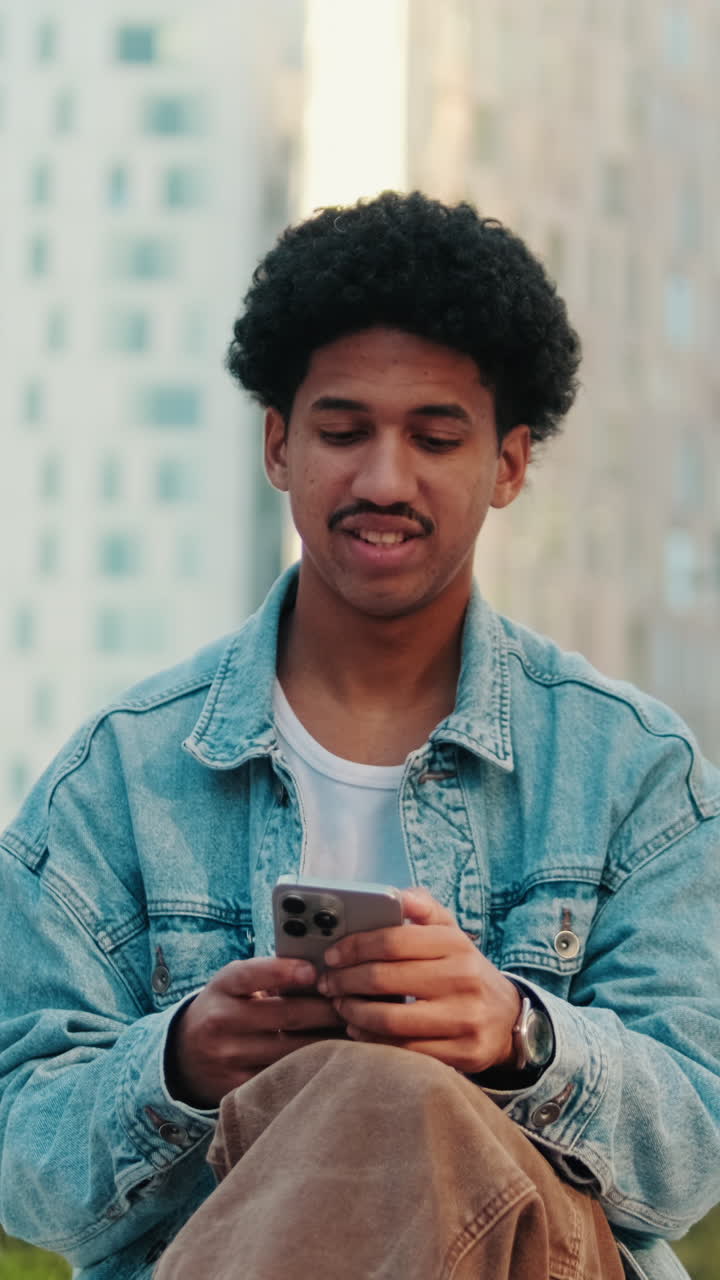 Giovane uomo con capelli ricci che sorride mentre usa lo smartphone all'aperto
