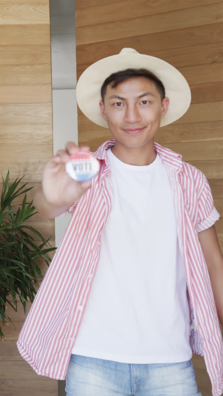 Vertical video: Asian male wearing straw hat is holding up a VOTE badge