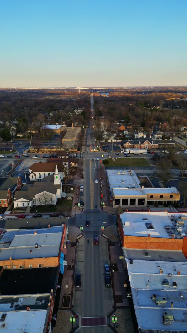 Una vista aerea cattura l'affascinante centro della città di Plainfield, Illinois, al tramonto, mostrando una vivace via principale fiancheggiata da edifici storici e un prominente campanile.