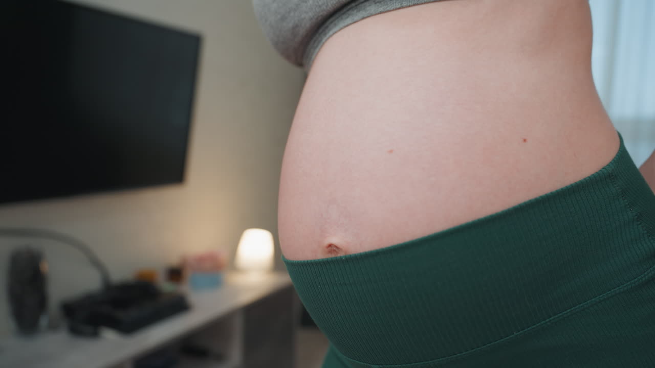 Primo piano del ventre di una donna incinta caucasica in un accogliente soggiorno. Mani sui fianchi, che indossa leggings verdi a vita alta, luce naturale soffusa dalla finestra, televisione e scrivania visibili, bagliore della lampada, calma intima.