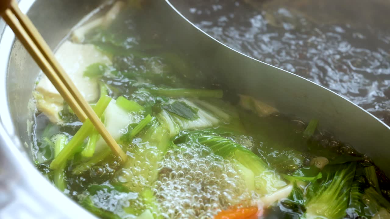Chopsticks stir leafy greens in boiling dual broth hotpot, close-up, natural daylight, minimal movement