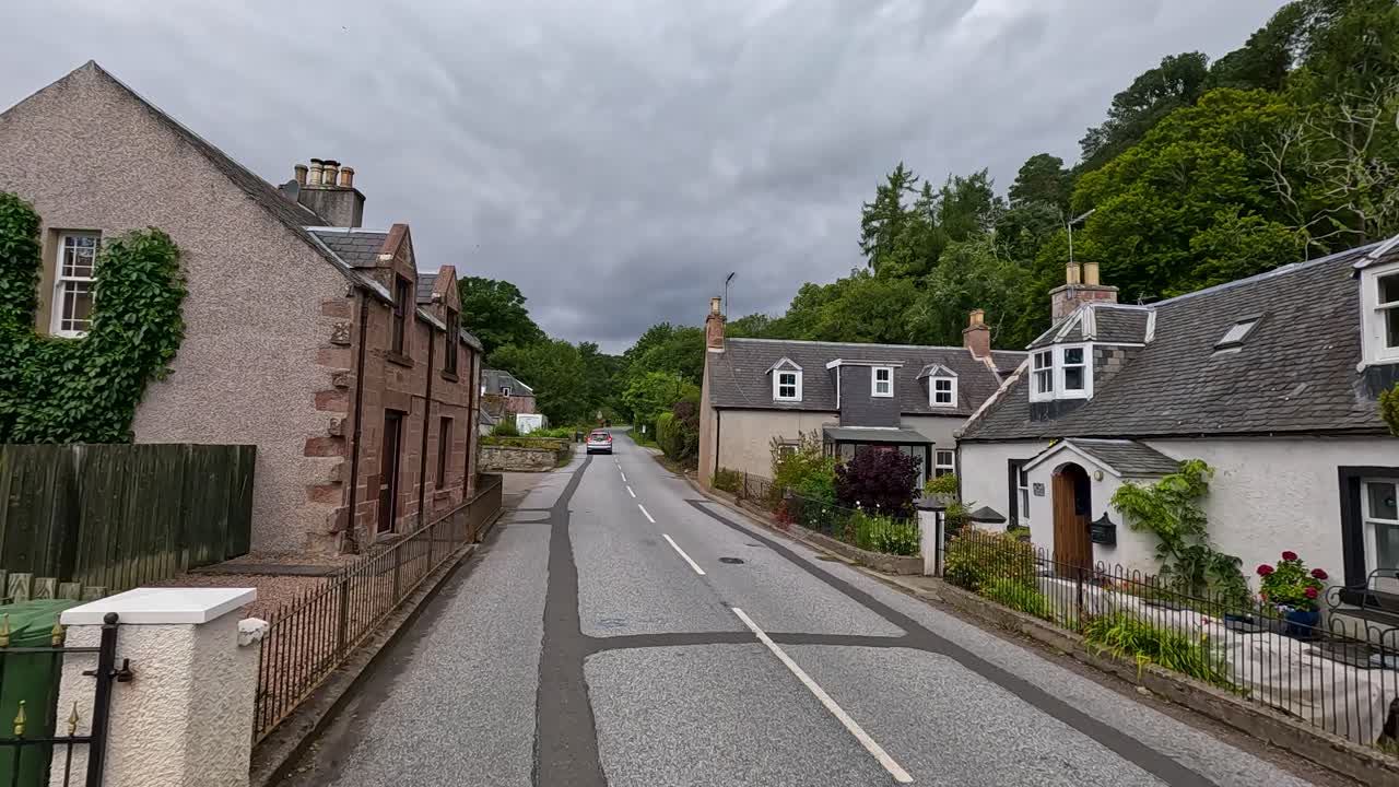 A vehicle moves along a narrow, empty street lined with traditional stone houses in a picturesque Scottish village under overcast daylight, captured in smooth forward motion