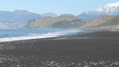 sea spray hangs in air before low hills and a mountain range on a mid-winter's day - black sand beach, kaikoura coastline