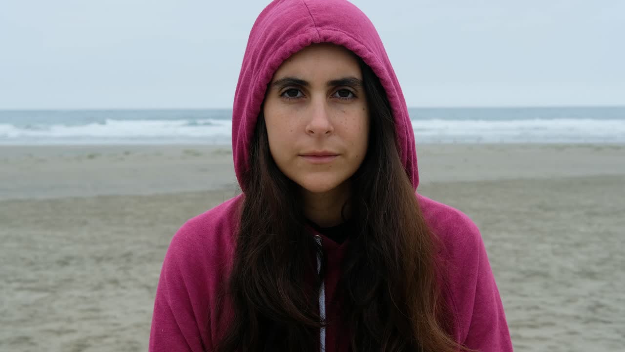 Ritratto ravvicinato di giovane donna bruna dall'aspetto casual che sorride con il cappuccio, mentre guarda la fotocamera in spiaggia, con l'oceano sullo sfondo.