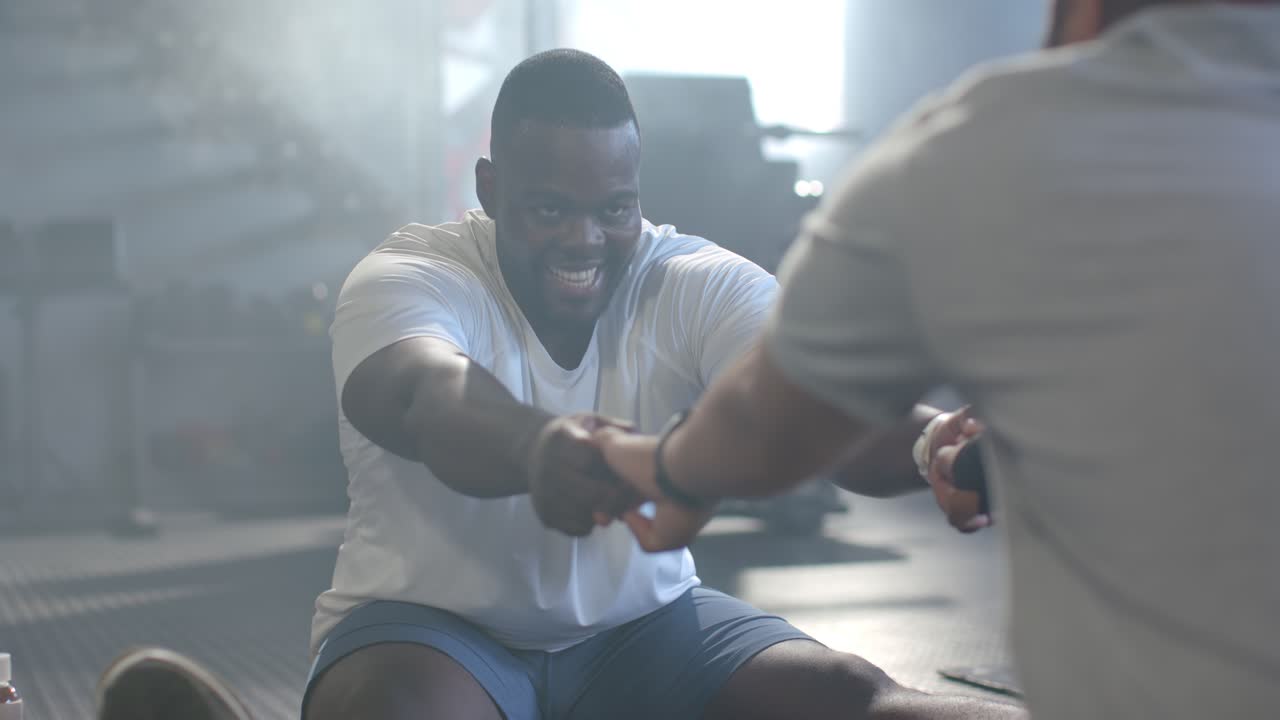 African American men, man in white tee pulling handle for strength while partner holding at gym