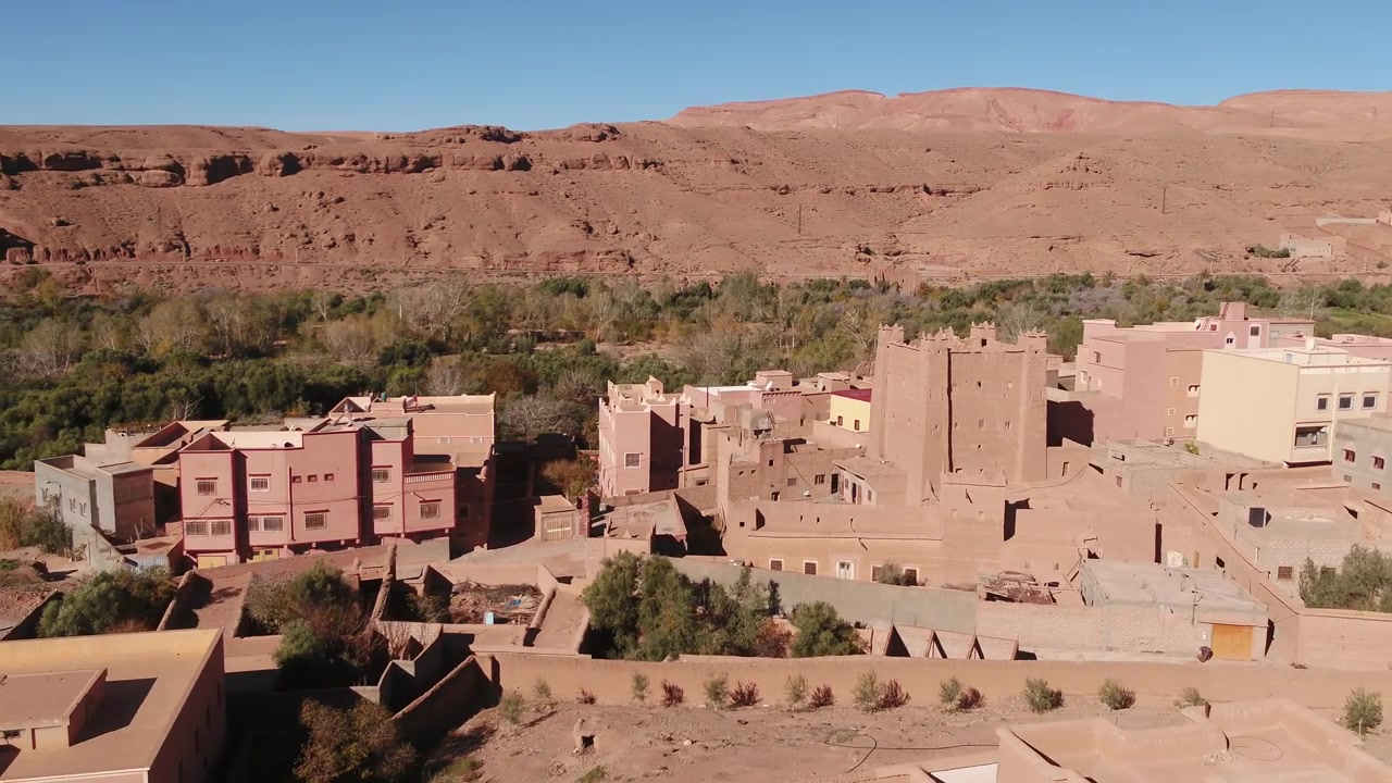 Ripresa aerea dall'alto che mostra una storica Kasbah in terra circondata da moderne case rurali e palmeti sotto aride montagne rosse a Tinghir, Marocco