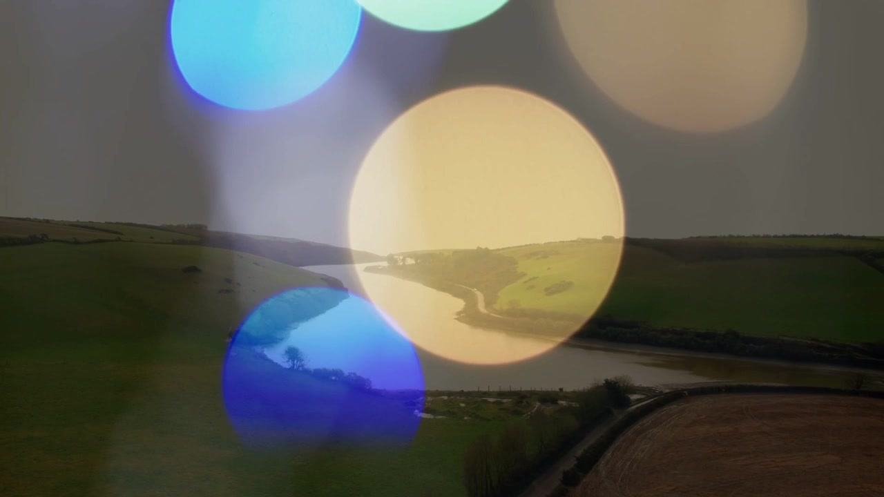Bokeh traslucido che appare, si sposta, si sovrappone e svanisce, rivelando un fiume serpeggiante e campi