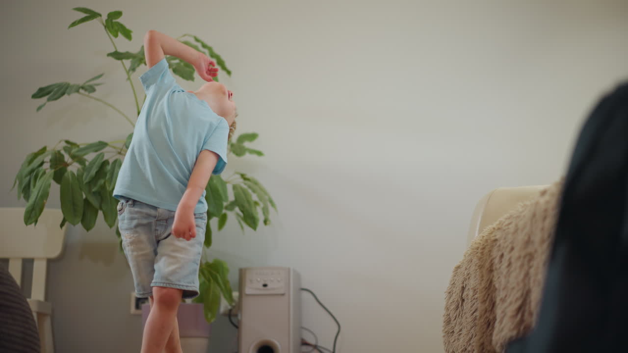 Happy little boy in blue shirt laughing with joy while standing near sofa in cozy living room, expressive playful face, carefree childhood moment, energy and innocence, bright mood, natural indoor lifestyle