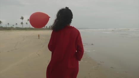 young indian brunette woman in a red dress with a red balloon in hand walking along the beach of the arabian sea in goa, india on a cloudy day