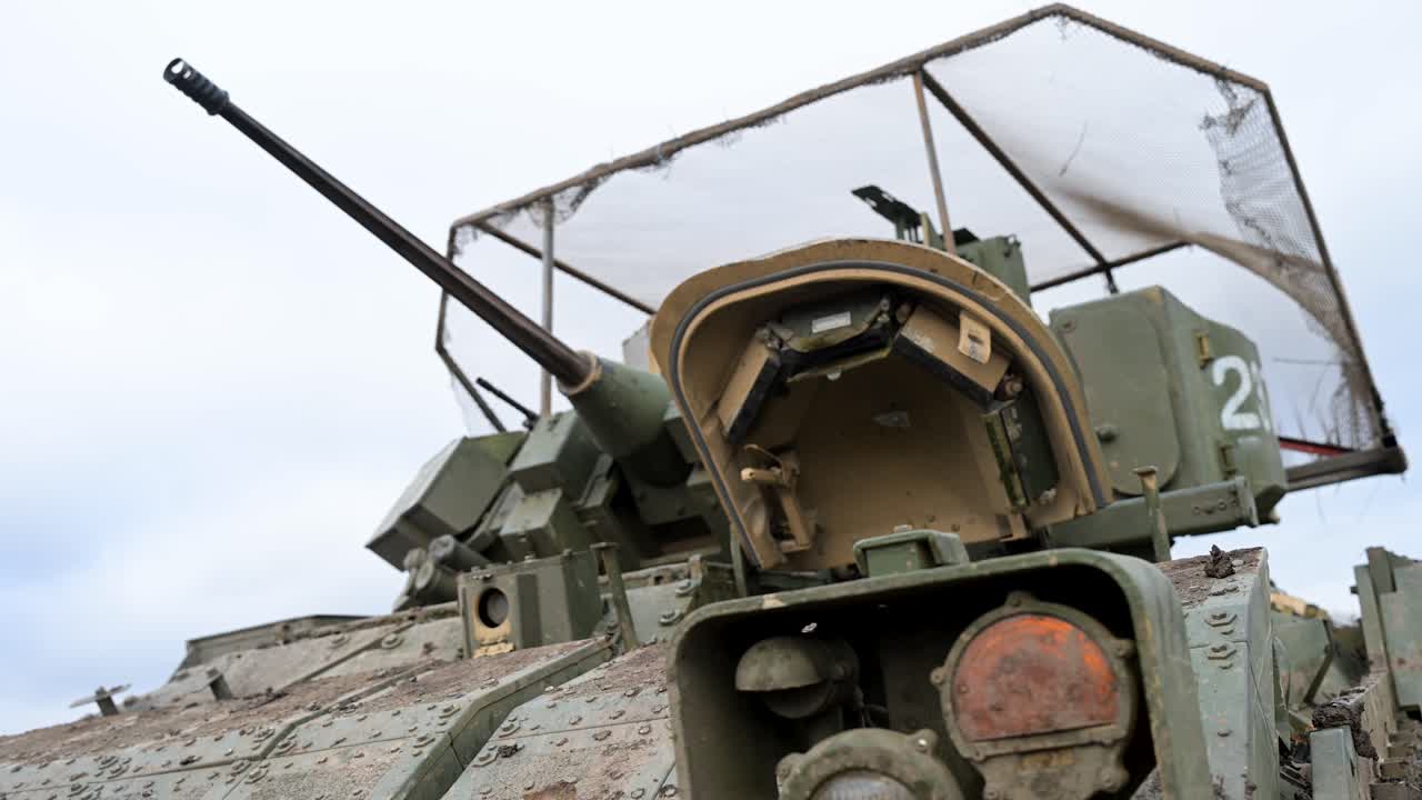A low-angle close-up of the 25mm Bushmaster autocannon on a US-made M2 Bradley IFV during a training exercise in Ukraine. A 'cope cage' is visible above the turret