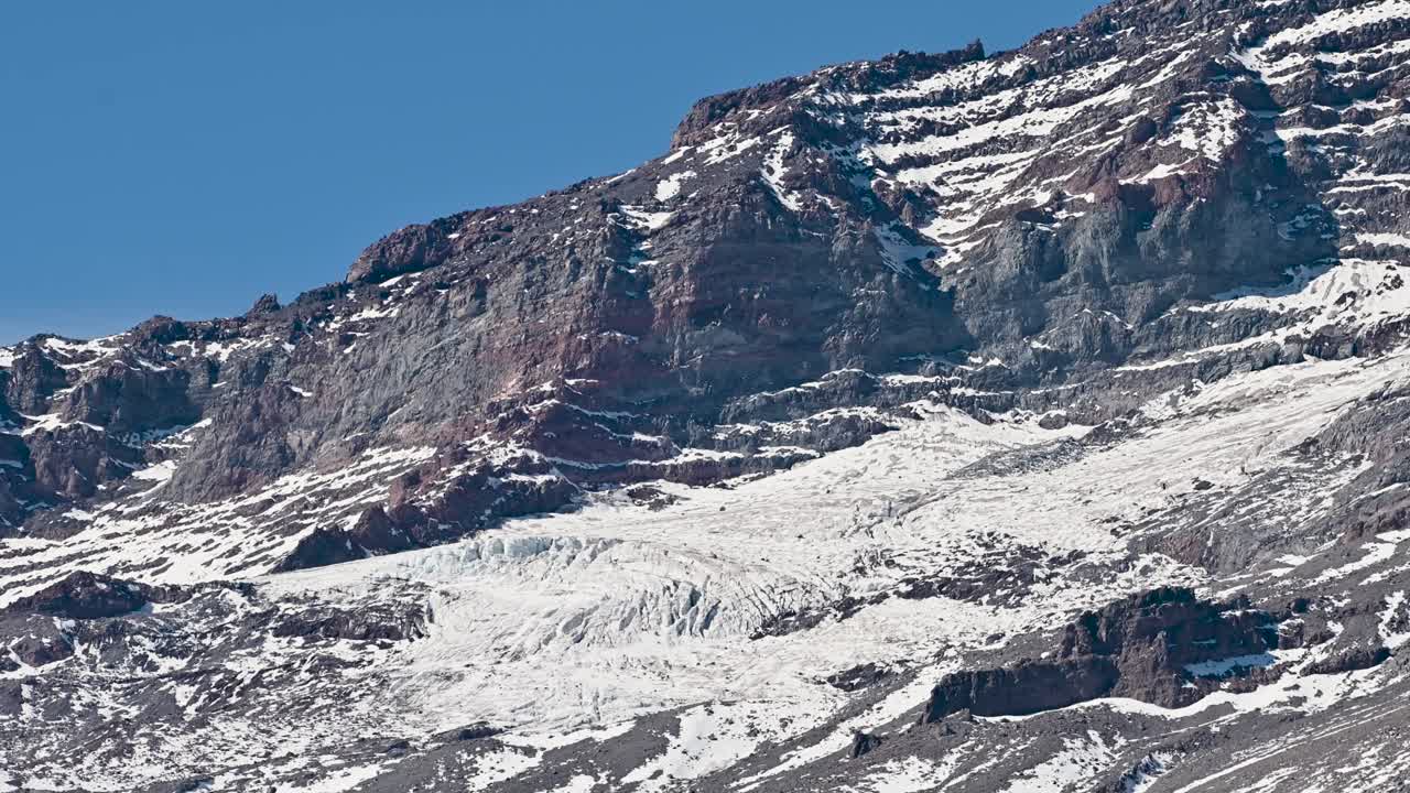 Snow-covered rocky mountain cliffs with exposed strata and glacial ice under a clear blue sky