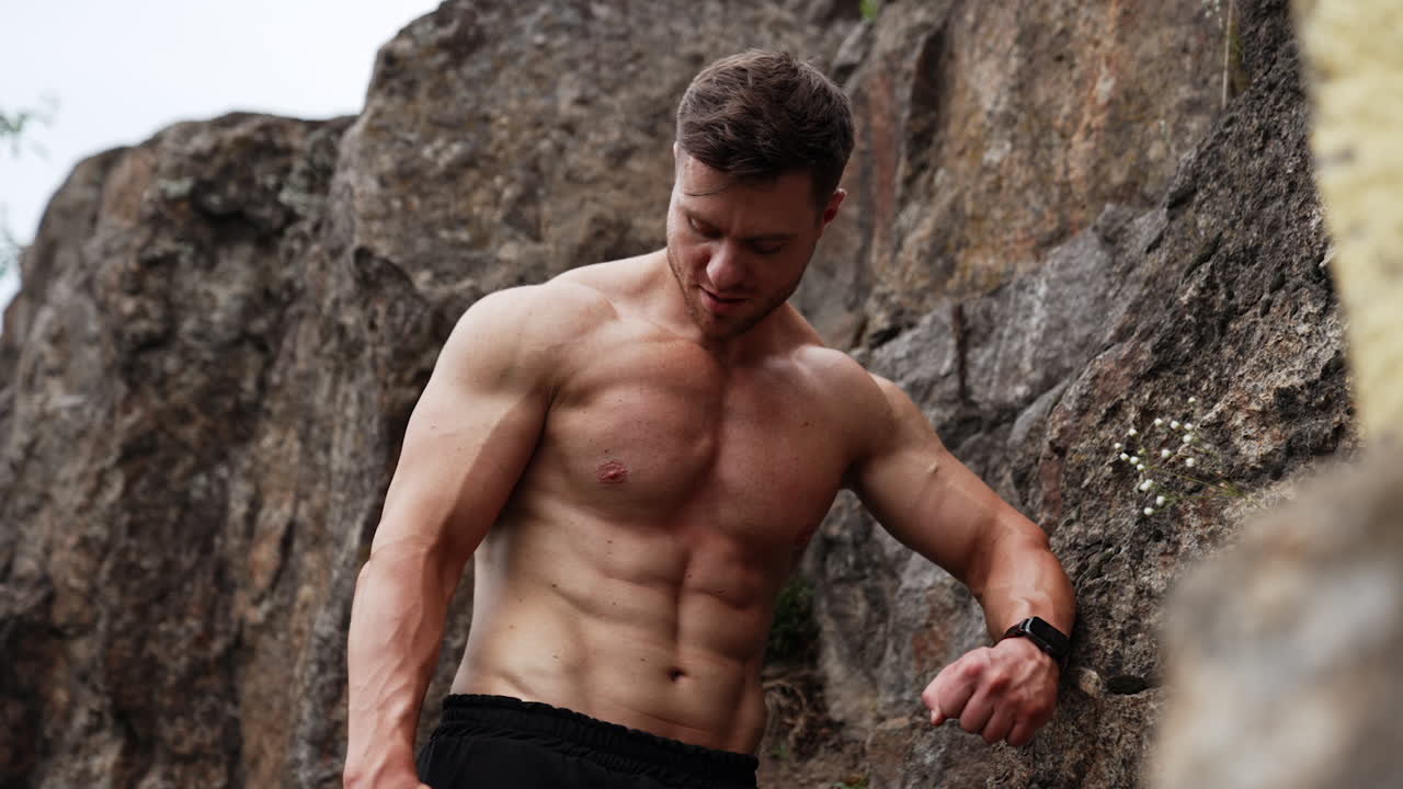 Athletic Caucasian man with naked torso stands at the rock. Sportsman taking a break during work out in the nature.