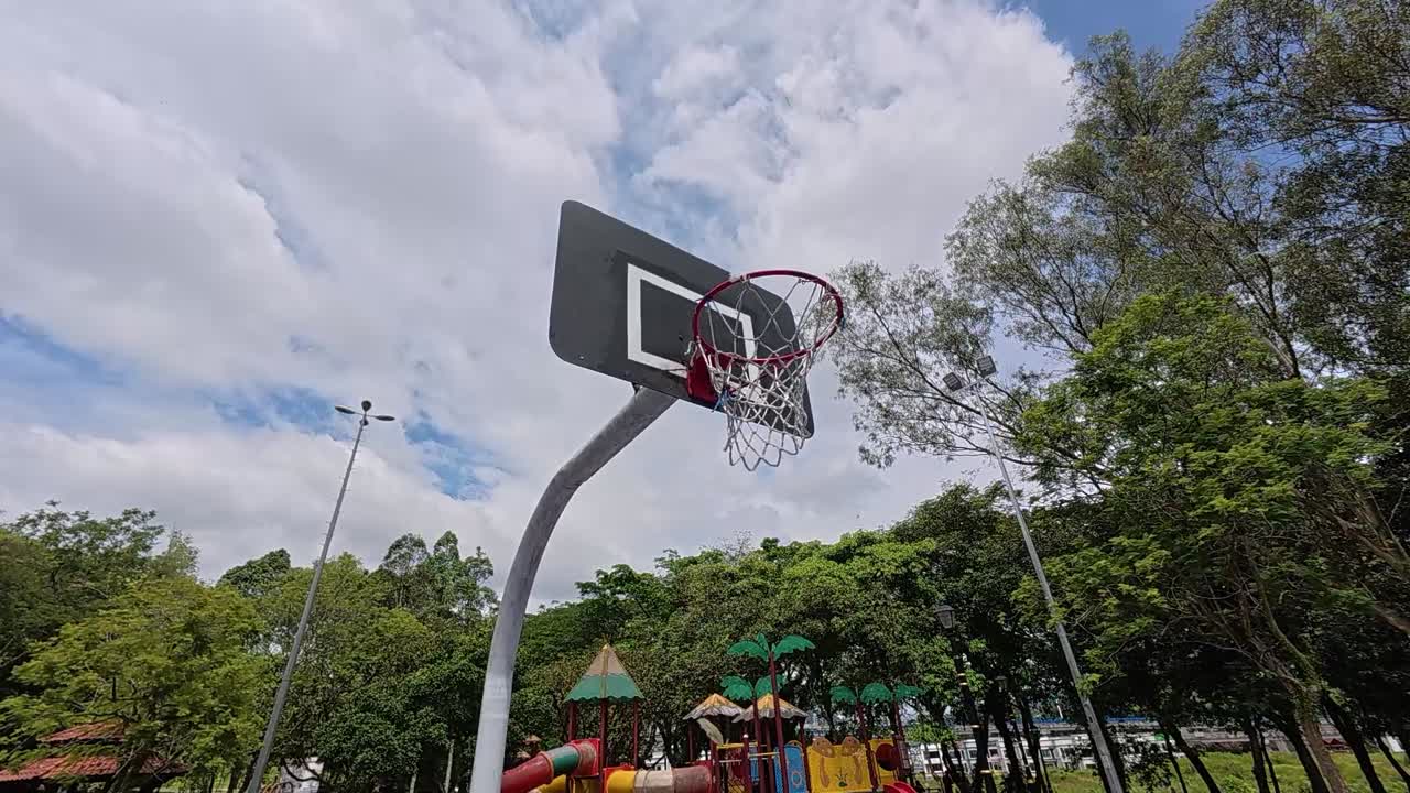 An empty basketball court in a tropical park, surrounded by lush green trees under a bright, sunny sky. Perfect weather.