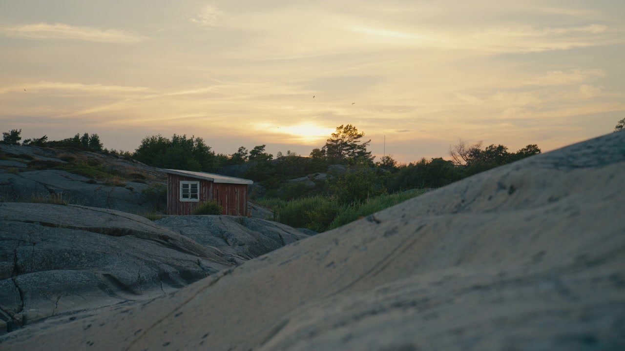Lille vejrbidt træbådhus beliggende blandt glatte granitklipper under solnedgang. Fredfyldt skandinavisk kystlandskab med varmt aftenlys, barsk kystlinje og rolig maritim atmosfære.