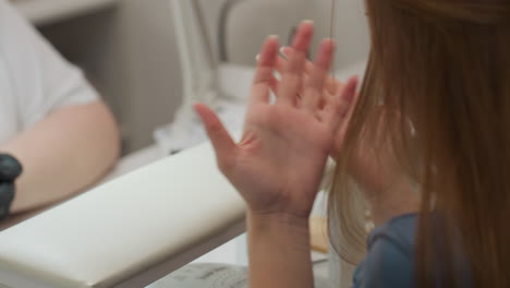 close up view of client raising hand to examine palm and nails before extending it toward nail technician holding electric file, preparing for manicure procedure in salon setting