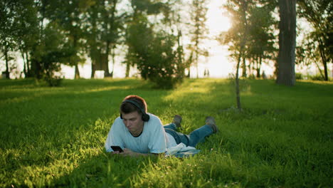 young man wearing black headphones lying on grass enjoying music and focusing on his phone in a scenic park setting, background features sunlight, greenery, and people walking in the distance