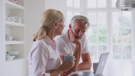 Mature-Couple-Sitting-At-Kitchen-Counter-Using-Laptop-To-Buy-Products-And-Services