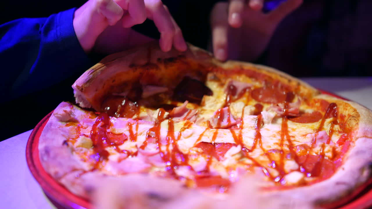Tearing and lifting a slice of pizza topped with pepperoni, ham, and drizzled sauce from the plate. The pizza's crispy crust and melted cheese create a mouthwatering display of a freshly served meal.