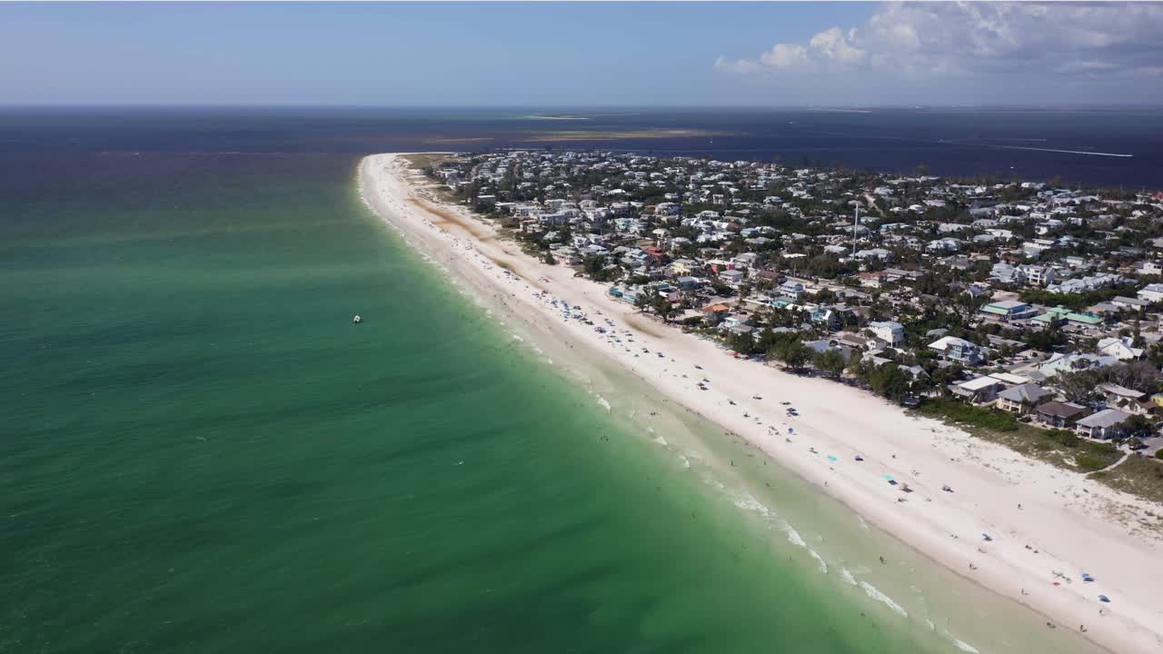 A sweeping view of Anna Maria Island captures the gentle curve of its white shoreline meeting clear emerald Gulf waters, with rows of coastal homes basking in bright afternoon light