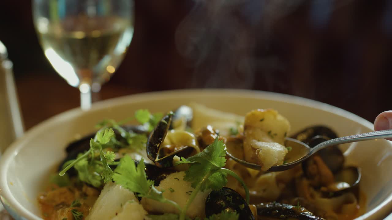 A hand uses a spoon to scoop steaming seafood chowder with mussels, squid, and herbs in a warmly lit restaurant setting, wine glass in background