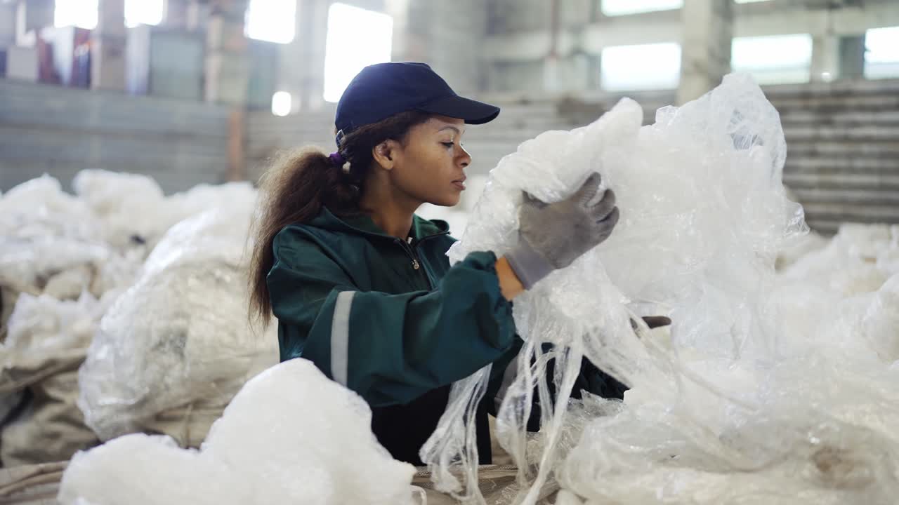 and African-American woman in a special uniform sorts polyethylene at a waste recycling plant. Processing of raw materials
