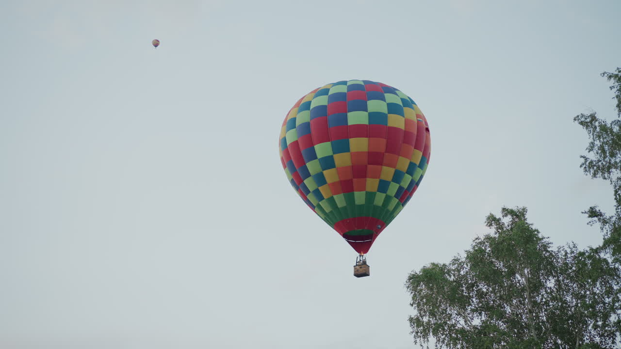 two vibrant checkered hot air balloons lifting passengers skyward with powerful burner flames against soft pastel dusk sky over expansive green field surrounded by dense forest