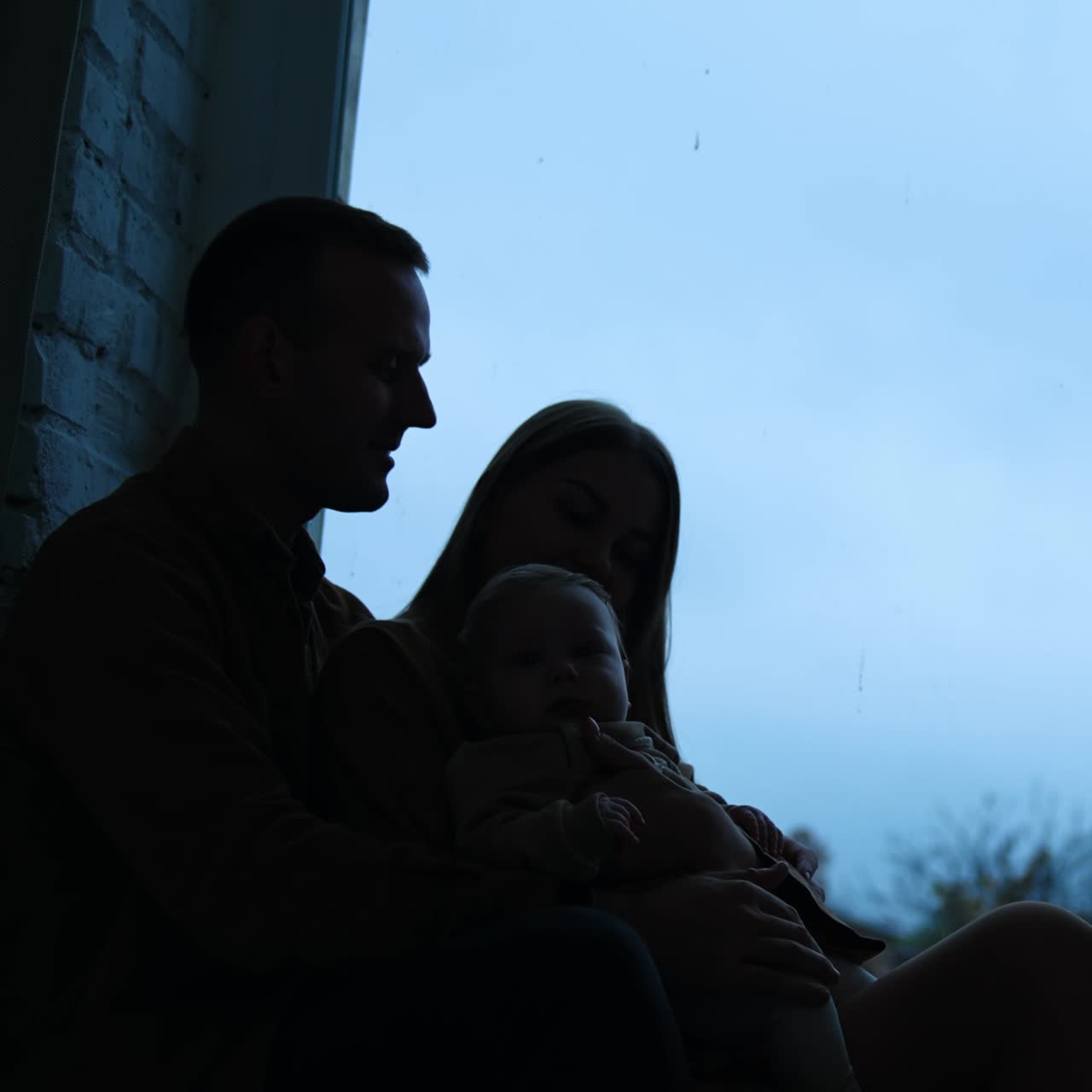 Famiglia di tre persone seduta sul davanzale della finestra di sera. Silhouette di genitori con un neonato
