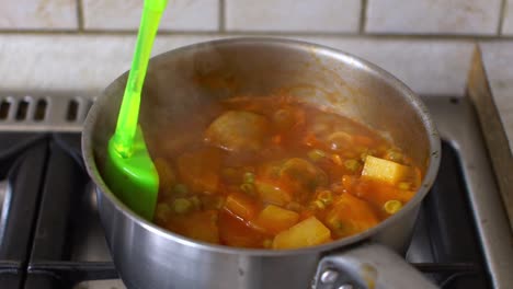 close up of boiled greek tomato pea stew with artichokes and potatoes , man stirring with a silicone spoon, slow motion