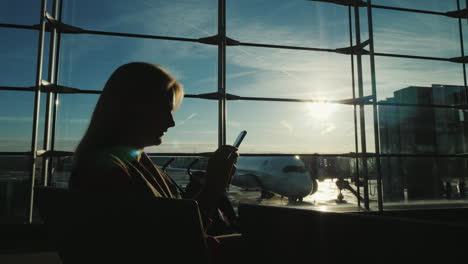 Silhouette-Einer-Frau,-Die-Mit-Einem-Smartphone-Am-Fenster-Im-Flughafenterminal-Sitzt-Sitting