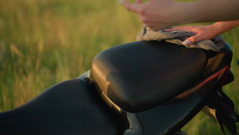 a close-up of a hand cleaning the seat of a motorcycle with a cloth in a grassy field, the person is carefully wiping down the bike