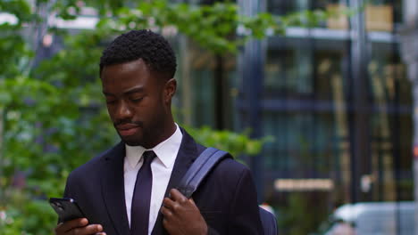 Young-Businessman-Wearing-Suit-Using-Mobile-Phone-Standing-Outside-Offices-In-The-Financial-District-Of-The-City-Of-London-UK