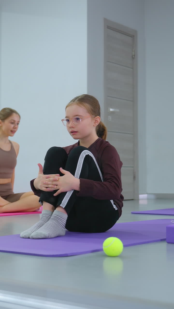 los niños en un gimnasio brillante realizan ejercicios en esteras, rodando hacia atrás con las manos juntas en las rodillas, bajo la vigilancia de un instructor visible en un espejo reflectante