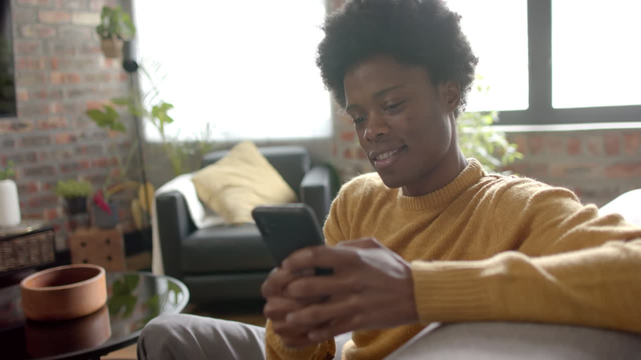 Happy african american man sitting on sofa using smartphone at home, slow motion