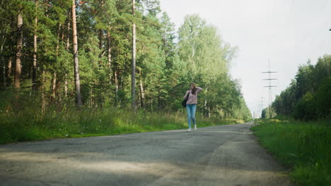 lady in pink hoodie walking on rural forest road with one hand in pocket and black shoulder bag, surrounded by dense green trees and overhead power lines stretching into distant