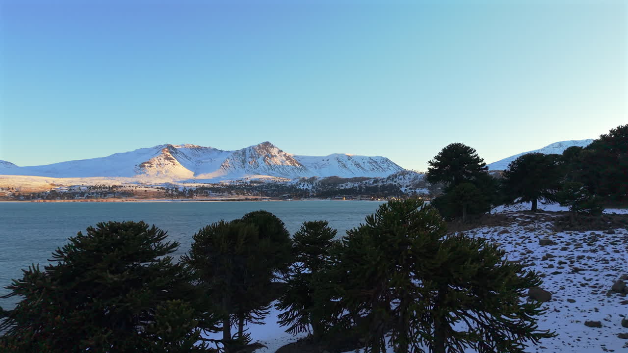 Aerial trucking shot revealing a beautiful sunset over the snow-covered Andes mountains and Lake Caviahue in Patagonia Argentina