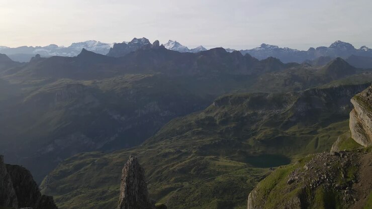 Aerial view of rugged mountain landscape with steep cliffs and green valleys, featuring the Gross Achslenstock in the distance, under a clear sky with distant snow-capped peaks