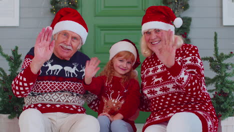 Senior-old-couple-grandparents-with-grandchild-girl-kid-waves-hand-hello,-hi-near-Christmas-house