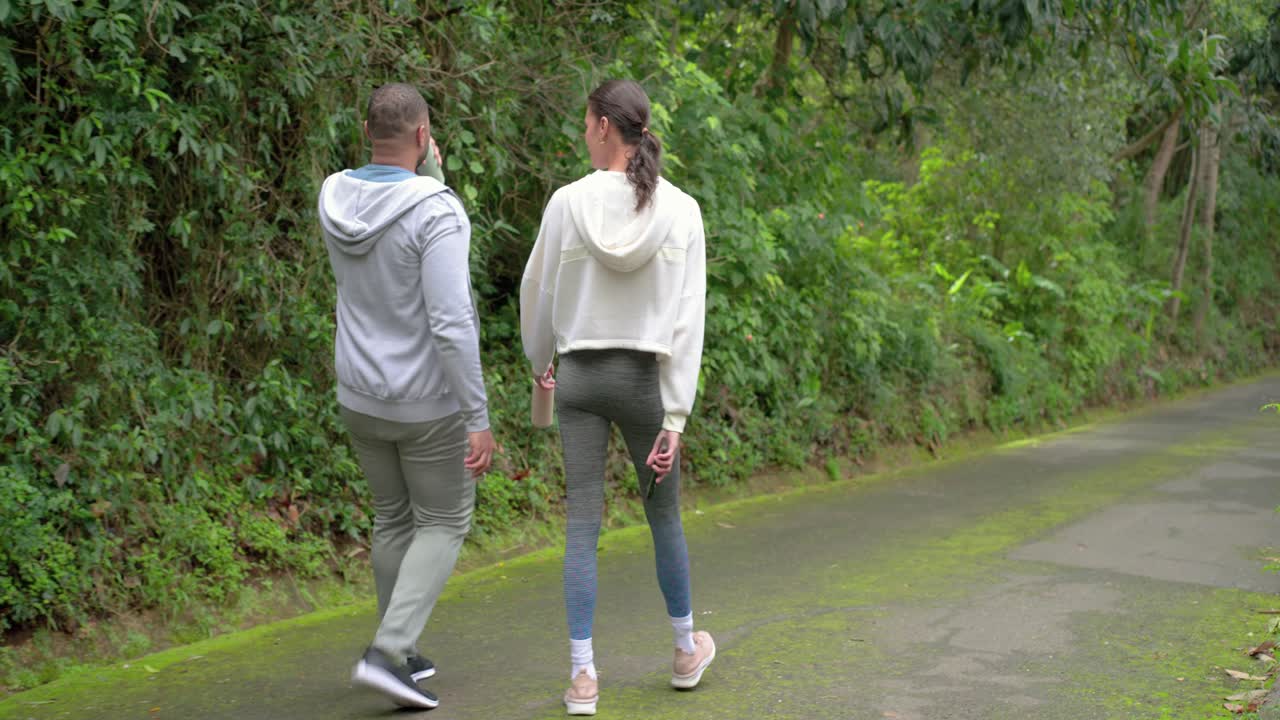 Diverse couple walking on trail holding water bottle and sipping tumbler for hydration and chatting