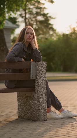 dama sentada en un banco al aire libre durante la hora dorada, descansando su cara en la mano, con expresión contemplativa mientras la luz del sol la ilumina suavemente, los alrededores incluyen caminos pavimentados, vegetación y edificios