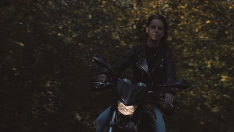 front view of beautiful european female motor biker wearing leather jacket on road with autumn leaf colored trees at amelisweerd forest on sunny day