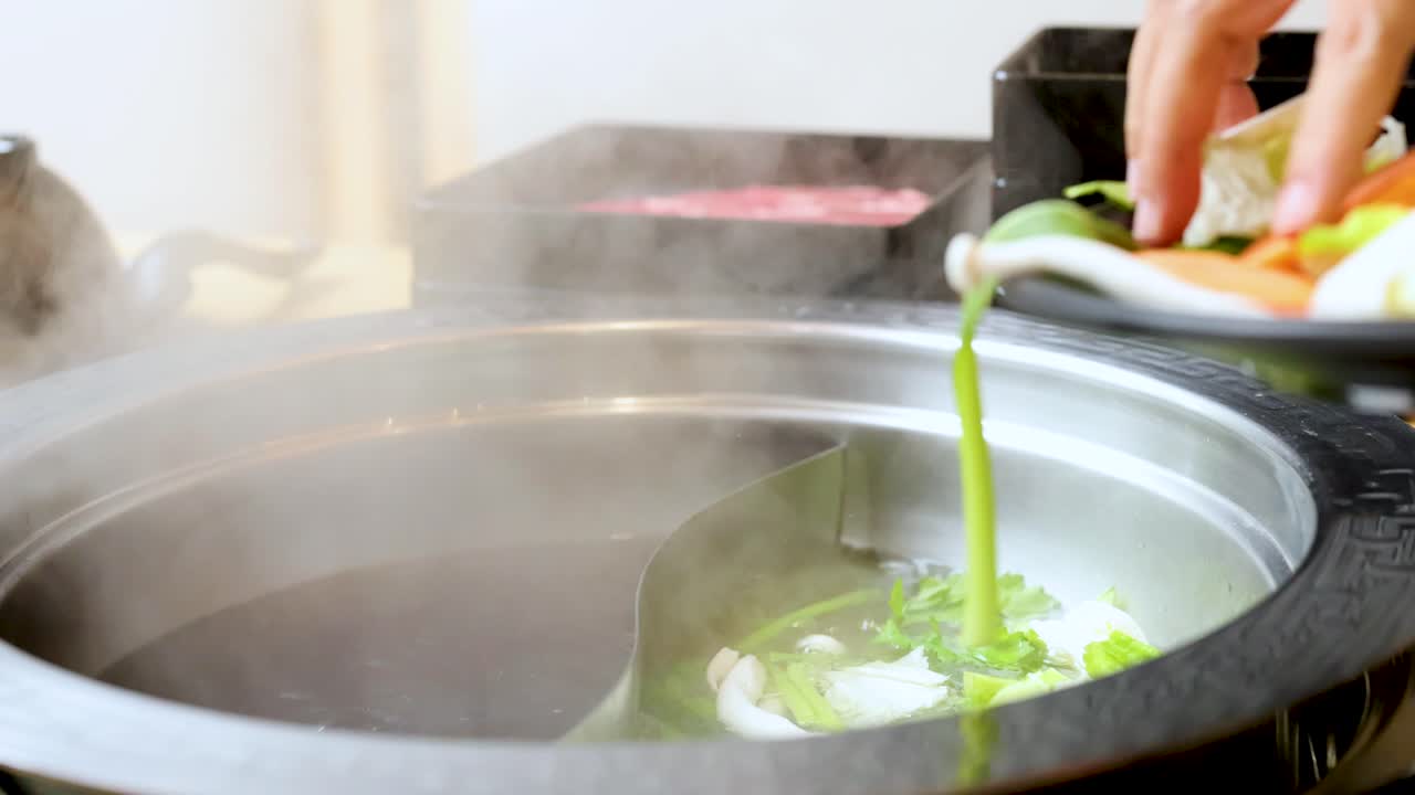 Hand drops assorted fresh vegetables into steaming dual broth hotpot, bright indoor lighting, close-up view